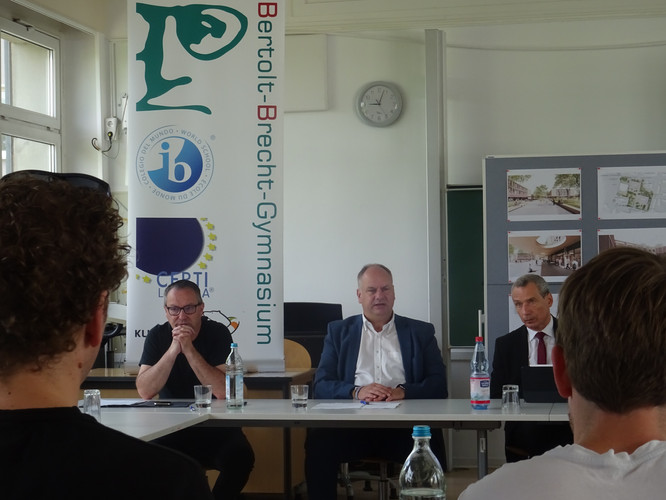 KI generiert: Das Bild zeigt drei Männer, die an einem Tisch in einem Klassenzimmer sitzen und vor ihnen eine Gruppe von Zuhörern. Im Hintergrund sieht man ein Banner mit der Aufschrift "Bertolt-Brecht-Gymnasium" und eine Pinnwand mit Architekturzeichnungen.