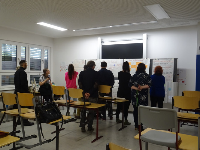 KI generiert: Die Hauptszene des Bildes zeigt eine Gruppe von Menschen in einem Klassenraum, die sich eine Präsentation oder Notizen an der Tafel ansehen. Umgedrehte und aufgestellte Stühle deuten darauf hin, dass das Treffen informell ist.