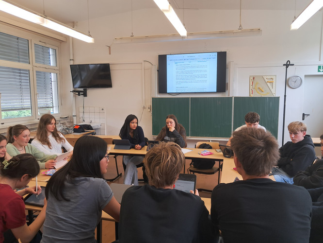 KI generiert: In einem Klassenzimmer sitzen Schülerinnen und Schüler in einem Halbkreis um Tische und schauen auf Tablets oder Notizen, während im Hintergrund ein Dokument auf einem Monitor angezeigt wird. Es sieht nach einer Gruppendiskussion oder einer Unterrichtsstunde aus.