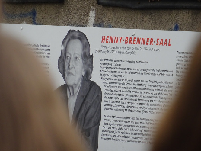 KI generiert: Das Bild zeigt eine Gedenktafel für den "Henny-Brenner-Saal" mit einer Schwarz-Weiß-Fotografie von Henny Brenner sowie einem Text, der ihr Leben und ihr Engagement zur Erinnerung an die Vergangenheit beschreibt. Teile des Bildes sind durch unscharfe Objekte im Vordergrund verdeckt.