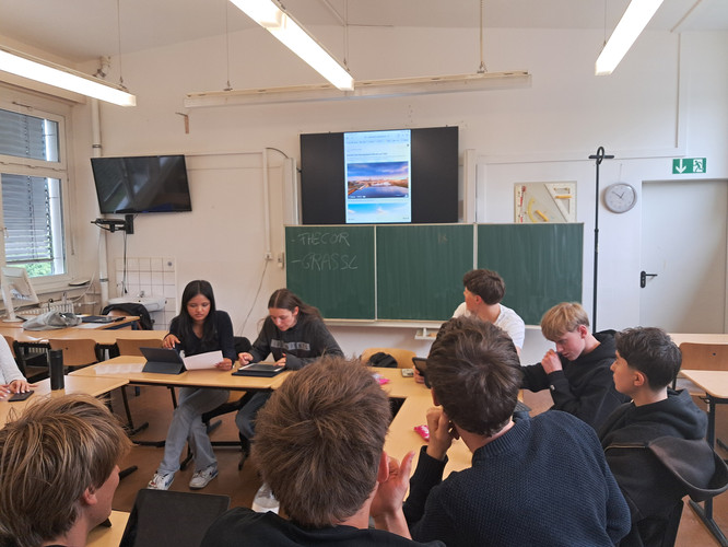 KI generiert: Eine Gruppe von Schülerinnen und Schülern sitzt in einem Klassenzimmer bei einer Präsentation oder Diskussion. Auf der Tafel sind einige Wörter geschrieben und ein Bild wird auf einem Monitor angezeigt.