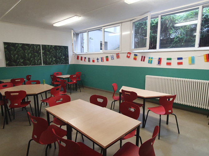 KI generiert: Das Bild zeigt einen Klassenraum mit mehreren Tischen und roten Stühlen. An der Wand sind kleine Fahnen verschiedener Länder aufgehängt.