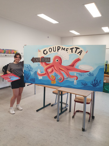 KI generiert: Eine Person steht neben einem bunt gestalteten Schild für eine Cafeteria namens "Gourmetta", auf dem ein Oktopus mit Kochutensilien zu sehen ist. Das Bild ist fröhlich und kreativ gestaltet, um die Cafeteria zu bewerben.