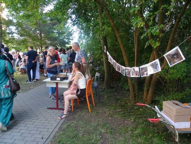 KI generiert: Das Bild zeigt eine Freiluftveranstaltung mit mehreren Menschen, die an einem Tisch versammelt sind und sich unterhalten. Im Hintergrund sind Fotos an einer Leine zwischen Bäumen aufgehängt.