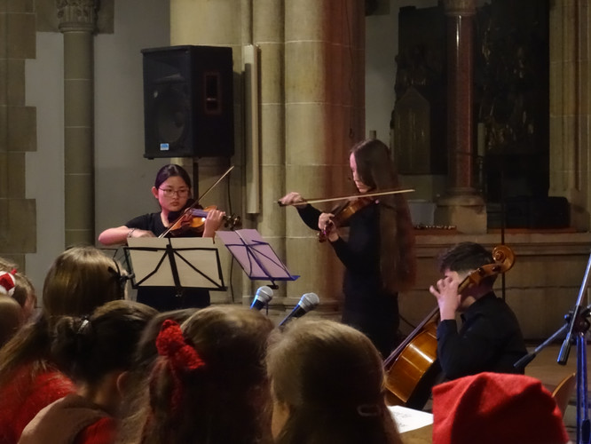 KI generiert: Das Bild zeigt ein Trio von Musikern in einer Aufführung, die Streichinstrumente spielen. Im Vordergrund sitzen Zuschauer, offenbar in weihnachtlicher Stimmung.