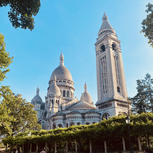 KI generiert: Das Bild zeigt die Basilika Sacré-Cœur in Paris bei sonnigem Wetter. Umgeben von Bäumen und mit klarem Himmel vermittelt es ein eindrucksvolles architektonisches Bild.