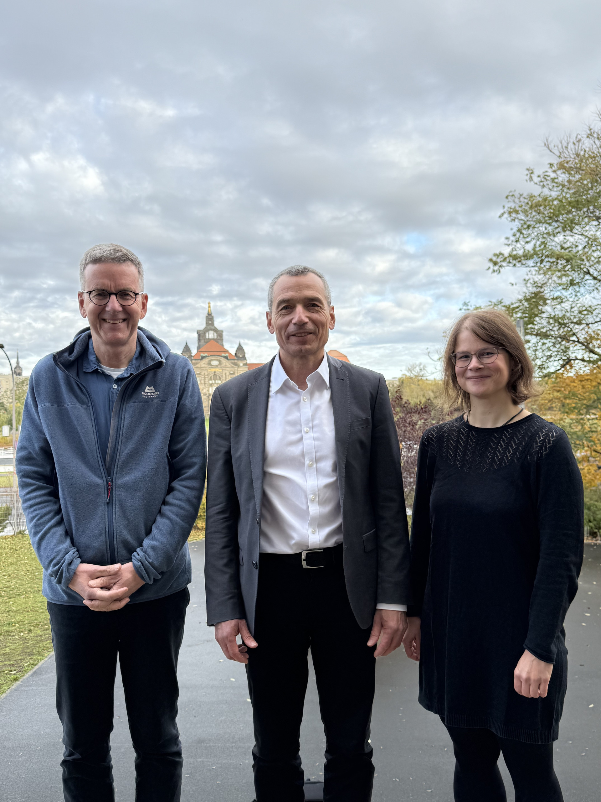 KI generiert: Das Bild zeigt drei Menschen, die in einer Außenumgebung nebeneinander stehen und in die Kamera lächeln. Im Hintergrund ist eine historische Gebäude mit Türmen sowie Wolken am Himmel zu sehen.