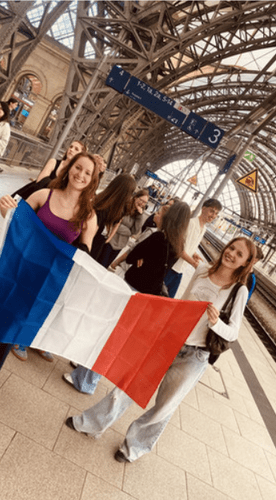 KI generiert: Das Bild zeigt eine Gruppe von jungen Menschen an einem Bahnhof, von denen zwei eine französische Flagge halten. Im Hintergrund sind Bahngleise und die Architektur des Bahnhofsgebäudes zu sehen.