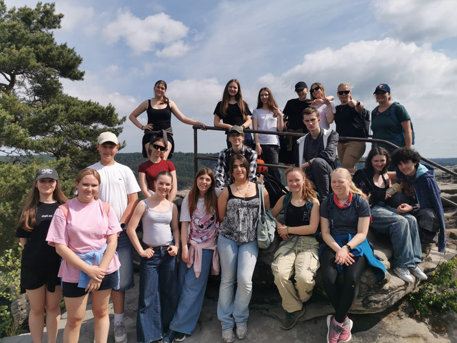 KI generiert: Eine Gruppe von Menschen steht gemeinsam auf einem Felsen in einer natürlichen Umgebung, offenbar auf einer Wanderung oder einem Ausflug. Der Himmel ist teilweise bewölkt, und im Hintergrund sind Bäume zu sehen.