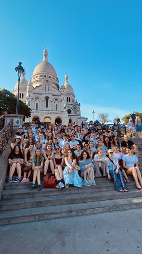 KI generiert: Eine große Gruppe von Menschen sitzt auf den Treppen vor der Basilika Sacré-Cœur in Paris. Im Hintergrund sieht man die beeindruckende Architektur der Kirche bei klarem Himmel.