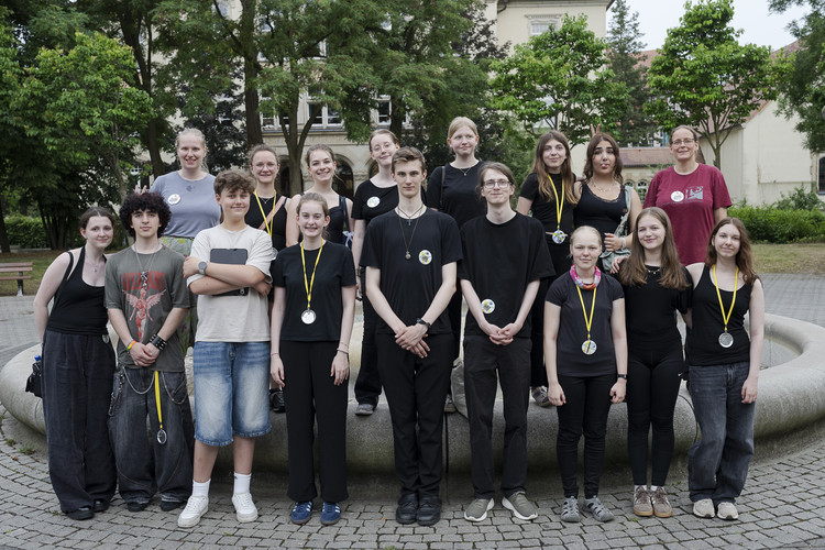 KI generiert: Eine Gruppe von jungen Menschen posiert draußen auf einem gepflasterten Platz für ein Gruppenfoto. Sie tragen Medaillen um den Hals und stehen in zwei Reihen vor einem Brunnen und Bäumen.
