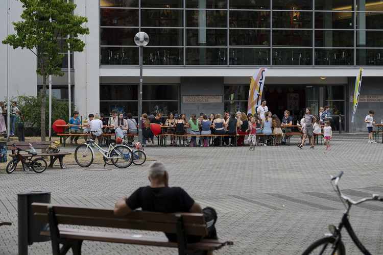 KI generiert: Auf dem Bild sind zahlreiche Menschen zu sehen, die sich um Tische vor einem modernen Gebäude versammelt haben, vermutlich zu einer Veranstaltung oder einem Fest. Im Vordergrund sitzt eine Person auf einer Bank, während mehrere Fahrräder herumstehen.