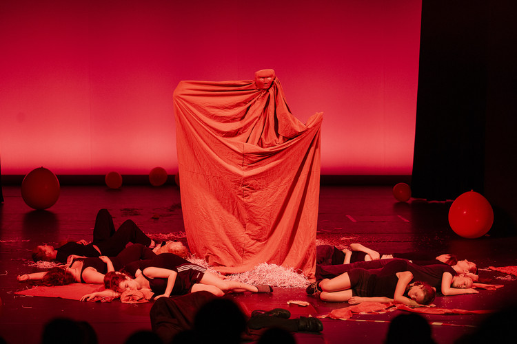 KI generiert: Das Bild zeigt eine Theateraufführung mit einer dramatischen roten Beleuchtung, bei der eine Person unter einem großen Tuch steht, während mehrere andere Darsteller auf dem Boden liegen. Große rote Luftballons sind im Hintergrund verteilt.