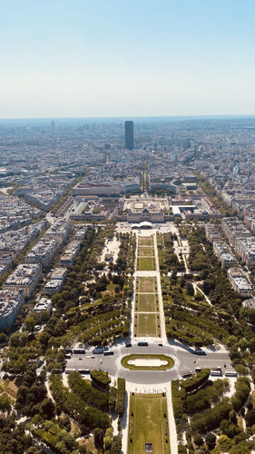 KI generiert: Das Bild zeigt einen Blick von oben auf den Champ de Mars, eine große Grünfläche, die sich in Richtung des Eiffelturms erstreckt, umgeben von symmetrisch angeordneten Gebäuden und Straßen in einer städtischen Landschaft. Im Hintergrund ragt ein markanter, schwarzer Turm hervor.