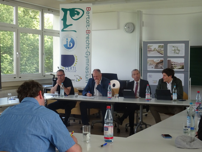 KI generiert: Das Bild zeigt eine Gruppe von Männern, die in einem Besprechungsraum an einem Konferenztisch sitzen. Im Hintergrund sind ein Banner des Bertolt-Brecht-Gymnasiums und Plakate mit Architekturplänen zu sehen.
