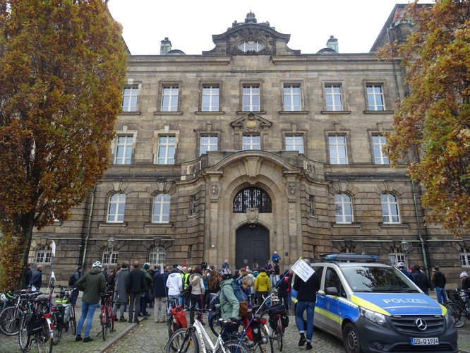 KI generiert: Das Bild zeigt eine Gruppe von Menschen, die sich vor einem großen, historischen Gebäude versammelt hat, das eine beeindruckende Fassade und einen großen Eingang besitzt. Vor dem Gebäude steht ein Polizeiwagen, was auf eine organisierte Veranstaltung hindeutet.