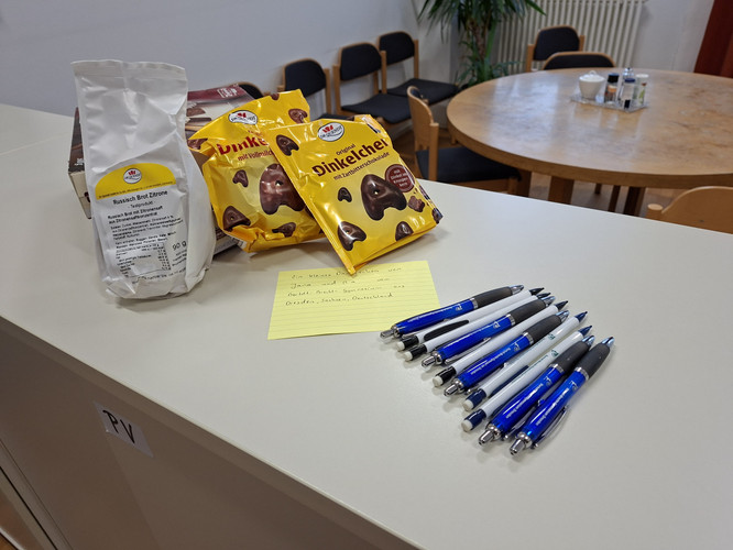 KI generiert: Das Bild zeigt eine Sammlung von Snacks und Stiften auf einem Tisch. Im Hintergrund sind Stühle um einen runden Tisch zu sehen.