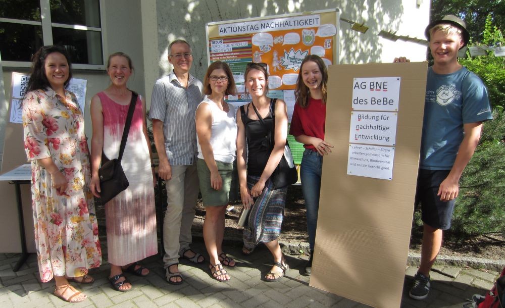 KI generiert: Eine Gruppe von Menschen steht vor einem Informationsplakat zum Thema Nachhaltigkeit und hält ein Schild mit der Aufschrift "Bildung für nachhaltige Entwicklung". Der Anlass scheint ein Aktionstag für Nachhaltigkeit zu sein.