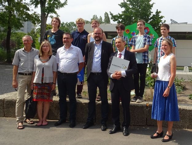 KI generiert: Eine Gruppe von Menschen posiert im Freien für ein Foto vor einer Steinumrandung, wobei einige von ihnen lächeln. Der Mann in der Mitte hält eine Mappe mit einem Logo in der Hand.