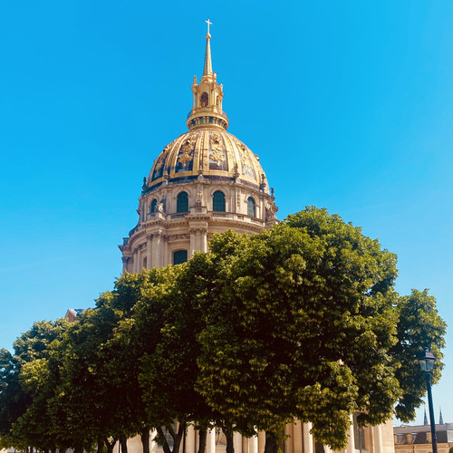 KI generiert: Das Bild zeigt die markante Kuppel eines Gebäudes mit goldenen Verzierungen, die über grünen Bäumen hervorragt. Der Himmel ist klar und blau, was die architektonischen Details der Struktur betont.