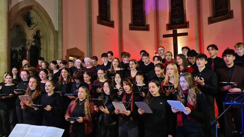 KI generiert: Das Bild zeigt einen Chor, der in einer Kirche singt. Die Chormitglieder sind in dunkler Kleidung gekleidet und einige halten Notenblätter oder Tablets in der Hand.