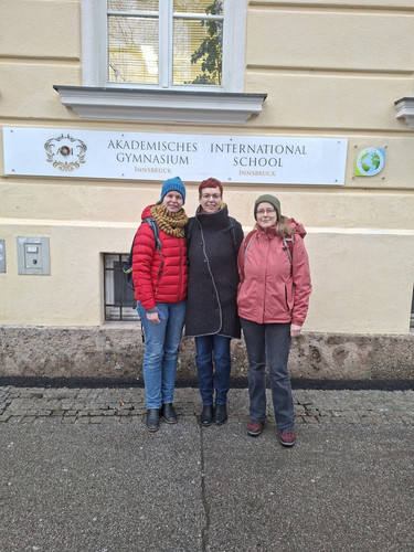 KI generiert: Drei Personen stehen vor einem Gebäude mit einem Schild, auf dem "Akademisches Gymnasium Innsbruck International School" steht. Alle tragen Winterkleidung und lächeln in die Kamera.