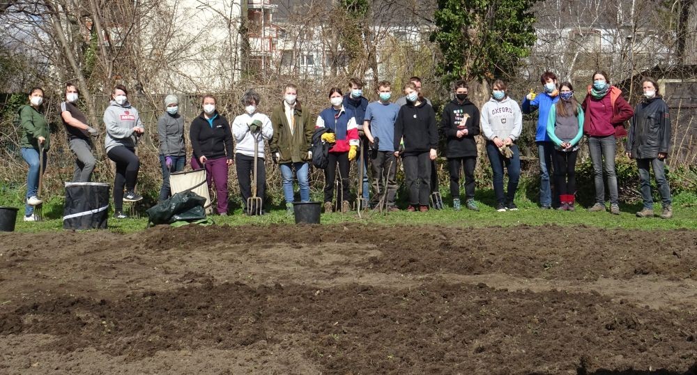 KI generiert: Das Bild zeigt eine Gruppe von Menschen, die in einem Garten stehen und teilweise Werkzeuge wie Spaten und Harken in den Händen halten. Sie scheinen bereit für Gartenarbeit zu sein und tragen überwiegend Masken.