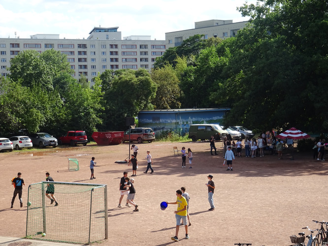KI generiert: Das Bild zeigt eine Gruppe von Menschen, die auf einem Sportplatz Fußball spielen. Im Hintergrund sind parkende Autos, Bäume und ein Wohngebäude zu sehen.