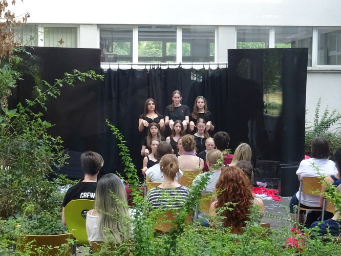 KI generiert: Das Bild zeigt eine Gruppe von Menschen, die in einem amphitheaterartigen Setup vor einem schwarzen Hintergrund posieren oder auftreten. Im Vordergrund sitzt ein Publikum, das der Szene zuschaut.