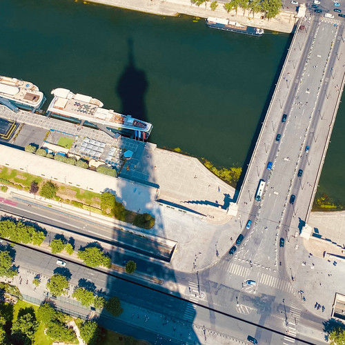KI generiert: Das Bild zeigt den Schatten des Eiffelturms, der über einen Fluss und eine Brücke fällt. Man sieht auch Autos auf der Brücke und Boote im Wasser.