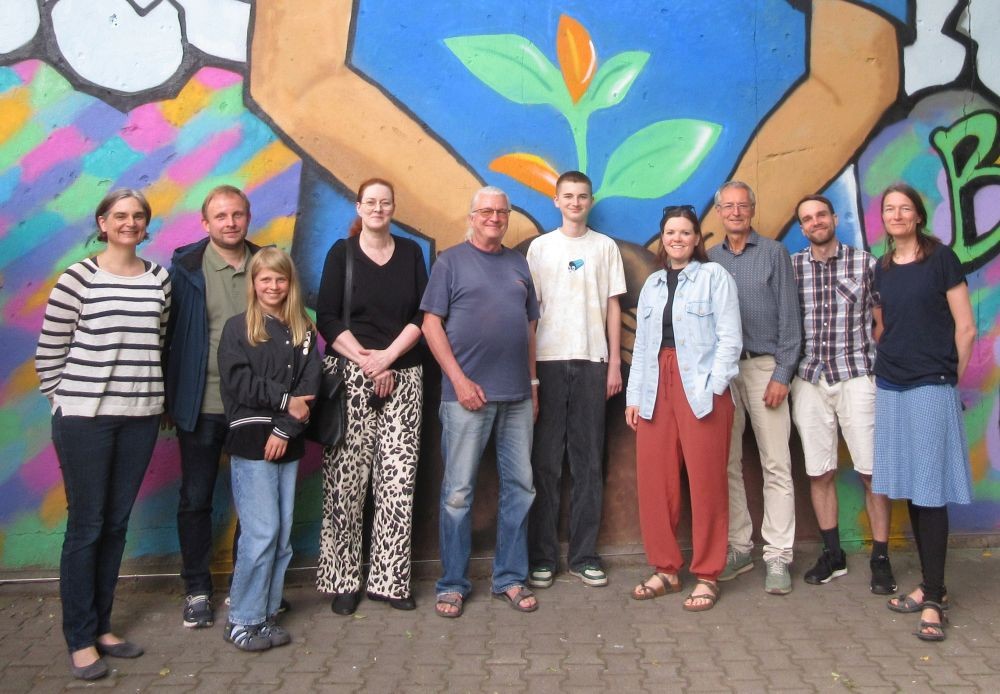 KI generiert: Das Bild zeigt eine Gruppe von Menschen, die vor einem bunten Wandgemälde stehen. Das Wandgemälde zeigt zwei Hände, die eine Pflanze halten.