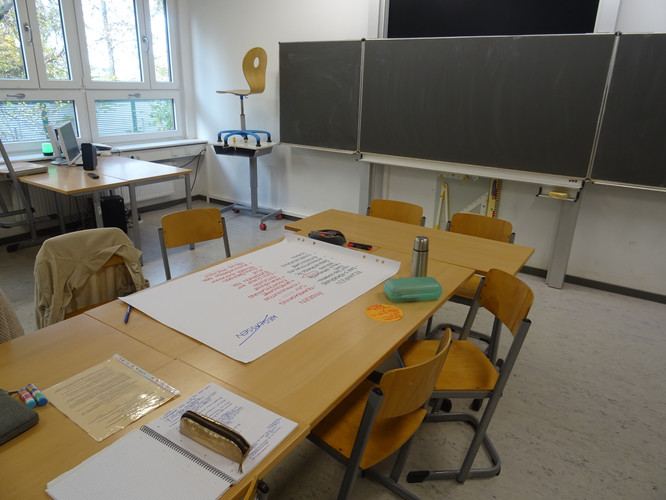 KI generiert: Das Bild zeigt einen Klassenraum mit Tischen und Stühlen, auf denen Schreibmaterialien und ein großes Blatt Papier mit Notizen liegen. Im Hintergrund sind eine weiße Tafel und ein Fenster zu sehen.