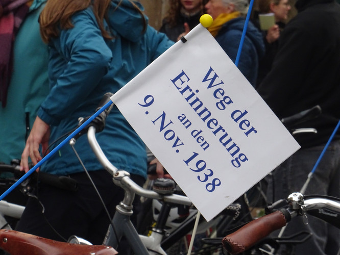 KI generiert: Das Bild zeigt ein Fahrrad, an dem ein Schild mit der Aufschrift "Weg der Erinnerung an den 9. Nov. 1938" befestigt ist, umgeben von Menschen. Es weist auf eine Gedenkveranstaltung zur Erinnerung an die Ereignisse der Kristallnacht hin.