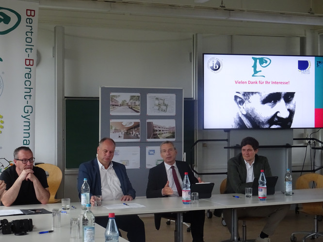 KI generiert: Das Bild zeigt eine Podiumsdiskussion oder Pressekonferenz mit vier Personen an einem Tisch in einem Klassenzimmer oder Konferenzraum, wobei im Hintergrund ein Bildschirm mit der Aufschrift "Vielen Dank für Ihr Interesse!" und dem Bild von Bertolt Brecht zu sehen ist. Es scheint sich um eine Veranstaltung im Bertolt-Brecht-Gymnasium zu handeln.