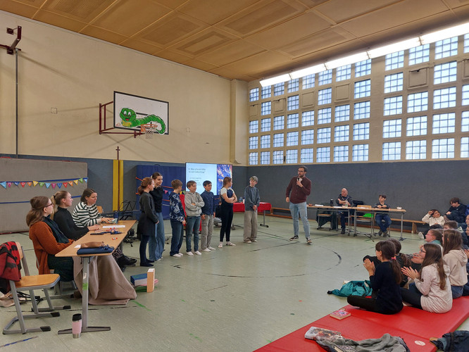 KI generiert: Das Bild zeigt eine Veranstaltung in einer Turnhalle, bei der einige Personen vorne stehen und ein Mann mit Mikrofon spricht, während andere an Tischen sitzen oder applaudieren. Es wirkt wie eine Präsentation oder eine Schulveranstaltung.