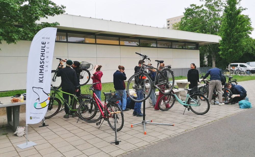 KI generiert: Das Bild zeigt eine Gruppe von Personen, die Fahrräder im Freien reparieren oder warten, neben einem Banner mit der Aufschrift "Klimaschule Sachsen". Im Hintergrund ist ein modernes Gebäude zu sehen.