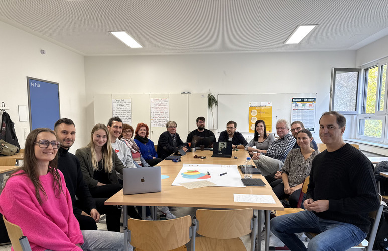 KI generiert: Eine Gruppe von Menschen sitzt an einem langen Tisch in einem Klassenzimmer, offenbar in einer Besprechung oder Schulung. Auf dem Tisch liegen Laptops und Papierbögen.