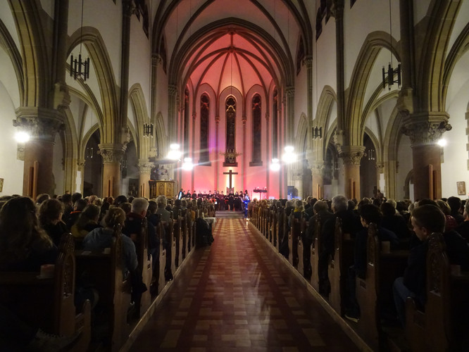 KI generiert: Das Bild zeigt den Innenraum einer Kirche mit einer großen Menschenmenge in den Bänken, die zum Altar blickt. Im Vordergrund führt ein Mittelgang zum Altarbereich, der in rotem Licht beleuchtet ist.