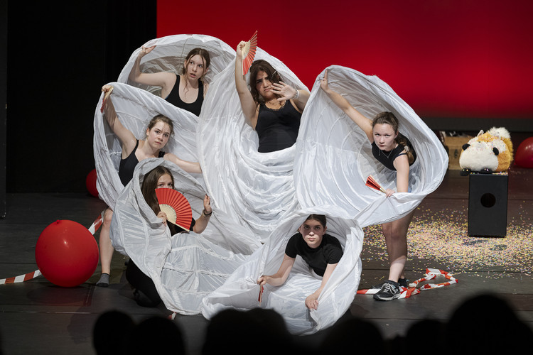 KI generiert: Das Bild zeigt eine Gruppe von Personen in einem Theaterstück, die in weißen Stoffbändern gewickelt sind und Fächer in den Händen halten. Im Hintergrund befinden sich rote Bälle und eine bunte Szenerie.