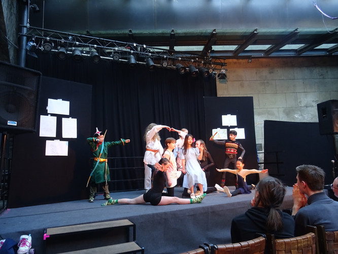 KI generiert: Das Bild zeigt eine Gruppe von Kindern auf einer kleinen Bühne, die in verschiedenen Kostümen eine Theateraufführung darbieten. Zuschauer im Vordergrund applaudieren der bunten Darbietung.