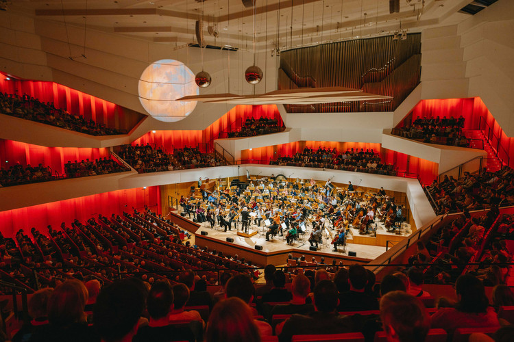 KI generiert: Das Bild zeigt ein symphonisches Orchester, das in einem modernen Konzertsaal vor einem großen Publikum spielt. Die warmen roten Lichter und die architektonischen Details des Saals schaffen eine einladende Atmosphäre.