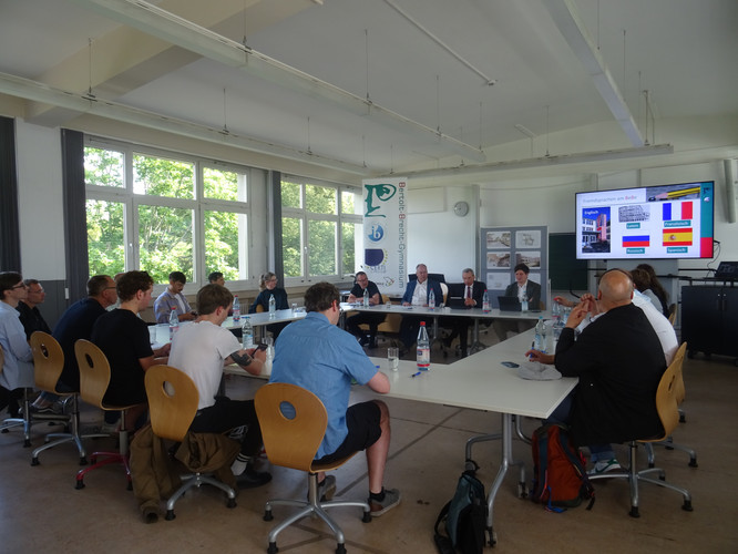 KI generiert: Das Bild zeigt eine Gruppe von Menschen, die an einem Konferenztisch in einem Raum mit moderner Ausstattung versammelt sind, wahrscheinlich zu einer formellen Besprechung oder Präsentation. Auf einem großen Bildschirm werden Informationen projiziert, und es sind mehrere teilnehmende Personen zu sehen.