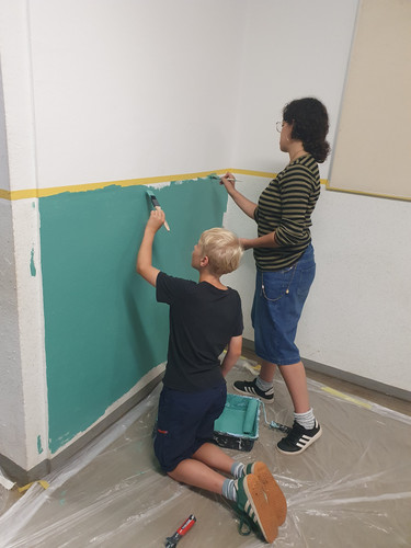 KI generiert: Zwei Personen malen gemeinsam eine Wand in einem Raum türkisfarben. Sie verwenden Pinsel, und der Boden ist mit Plastikfolie abgedeckt.