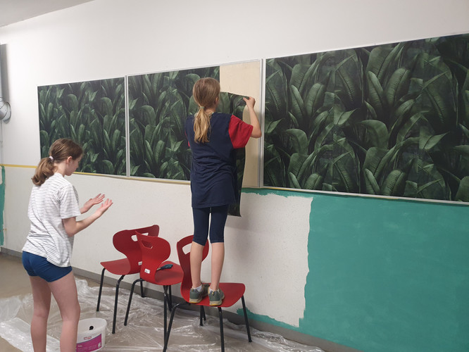 KI generiert: Zwei junge Personen bringen gemeinsam eine Fototapete mit Pflanzenmotiven an einer Wand an. Eine Person steht auf einem Stuhl, während die andere daneben steht und assistiert.