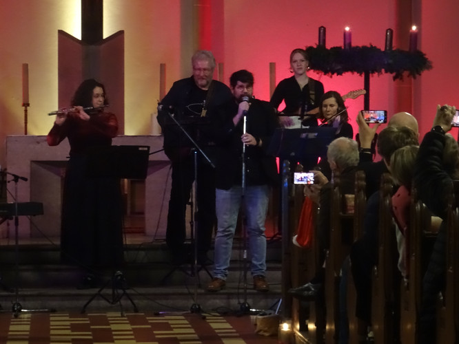 KI generiert: Das Bild zeigt eine musikalische Darbietung in einer Kirche, bei der mehrere Musiker auf einer Bühne spielen, während Zuschauer die Szene mit ihren Handys festhalten. Die Beleuchtung ist warm und eine Adventskranzdekoration ist im Hintergrund sichtbar.