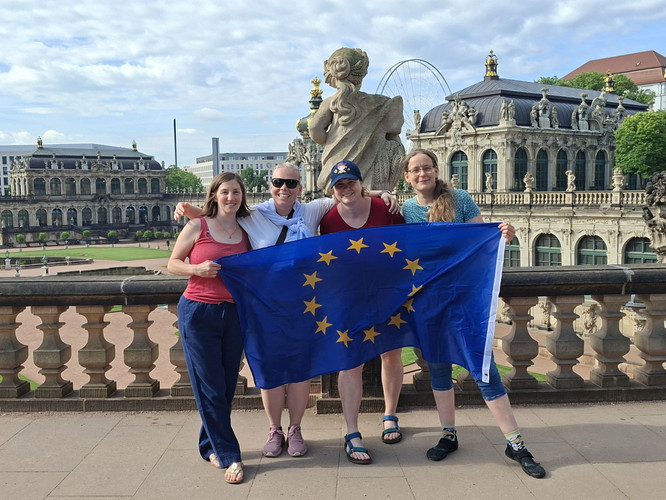 KI generiert: Vier Personen posieren lächelnd mit einer Europaflagge vor einer historischen Gebäudeanlage. Der Hintergrund zeigt eine barocke Architektur.