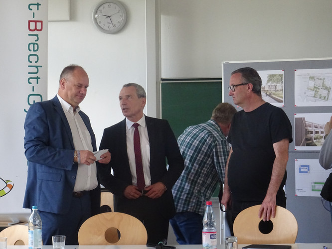 KI generiert: Das Bild zeigt eine Gruppe von Männern in einem Seminarraum, die in einer Diskussion vertieft sind. Im Hintergrund sind Plakate mit Architekturplänen und eine Tafel sichtbar.