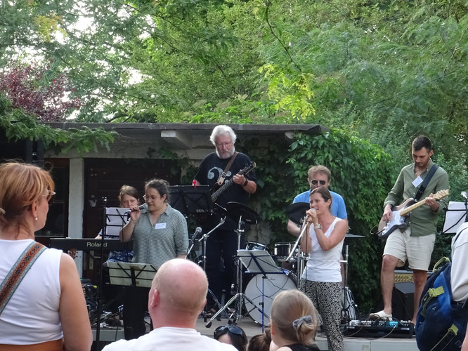 KI generiert: Das Bild zeigt eine Band, die im Freien auf einer Bühne auftritt, umgeben von grüner Vegetation. Mehrere Personen spielen Instrumente und singen, während Publikum im Vordergrund zuhört.