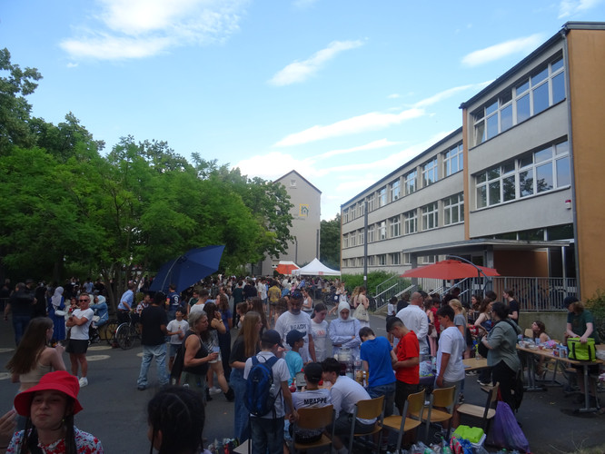 KI generiert: Das Bild zeigt eine lebendige Veranstaltung auf einem Schulhof, bei der viele Menschen zusammenkommen. Im Vordergrund sind Tische mit Aktivitäten oder Verkaufsständen zu sehen, während das Schulgebäude im Hintergrund steht.