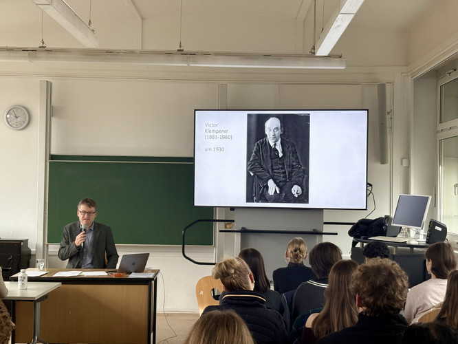 KI generiert: Ein Dozent hält einen Vortrag in einem Klassenraum, während im Hintergrund ein Bildschirm ein Bild von Victor Klemperer und seinen Lebensdaten zeigt. Vor ihm sitzen mehrere Zuhörer, die dem Vortrag folgen.
